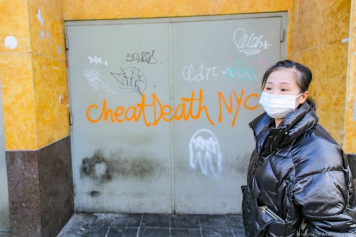 A photo of a woman wearing a surgical mask in front of graffiti that reads "Cheat Death NYC"